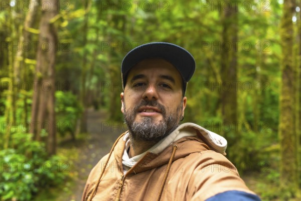 Tourist in a cap and light brown jacket taking a selfie while walking along a lush rainforest trail in ucluelet, british columbia, surrounded by natural beauty