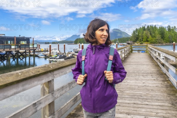 Female tourist wearing a purple jacket and carrying a backpack walking along a wooden pier in tofino, british columbia, enjoying the scenic view of moored boats and lush islands