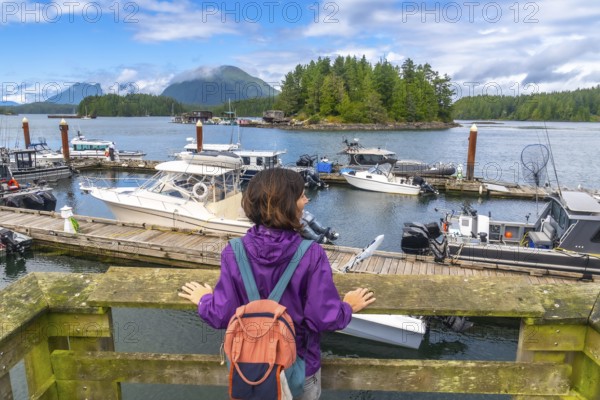 Female tourist with backpack contemplating moored boats in tofino harbor, vancouver island, british columbia, canada, during a sunny summer day with clouds and mountains in the background
