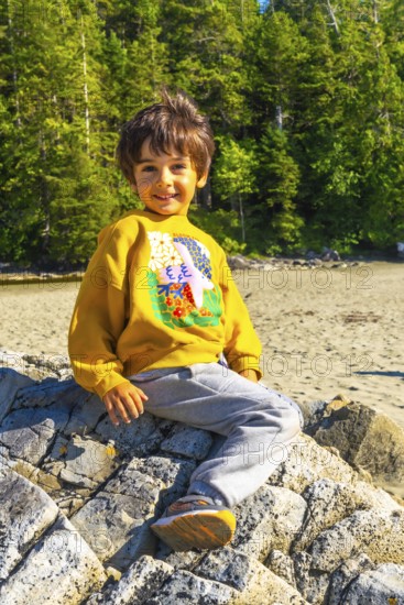 Happy child enjoying summer vacation, sitting on a rock at the beach in tofino, vancouver island, british columbia, canada, surrounded by lush green forest