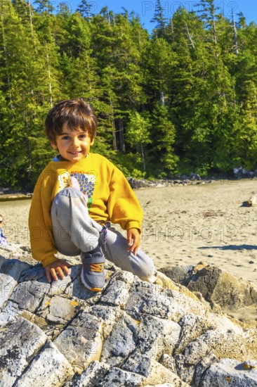Young boy sitting on a large rock at a beach in tofino, british columbia, soaking up the summer sun and enjoying the stunning natural scenery and vibrant surroundings