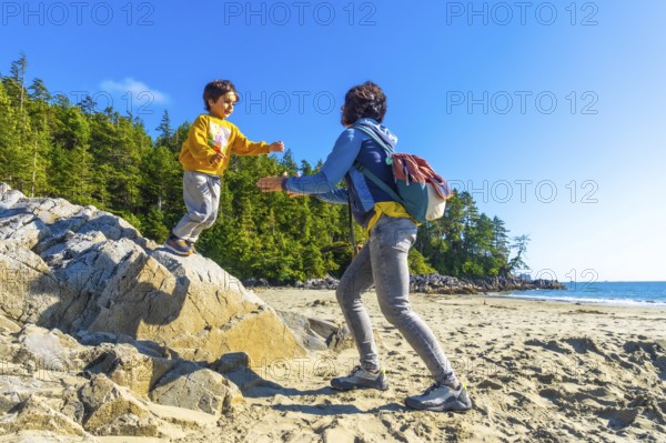 On a sunny summer day in tofino, british columbia, a mother extends her hands to help her child jump from a large rock on a sandy beach, surrounded by lush trees and the pacific ocean