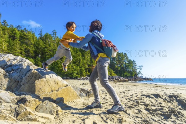 Mother catching her son as he joyfully jumps from a rock on a sandy beach, surrounded by lush green trees and a vibrant blue sky in tofino, vancouver island, canada