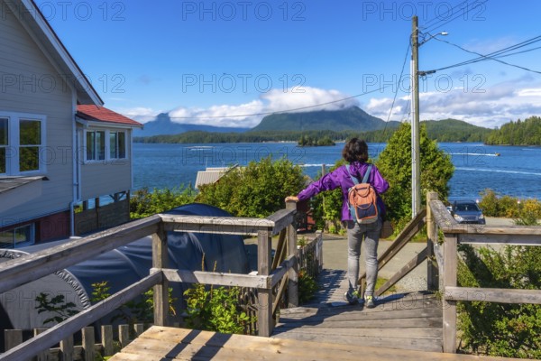 Female tourist with backpack walking down wooden stairs enjoying scenic view of tofino inlet, mountains, boats, and houses in tofino, vancouver island, british columbia, canada, during summer