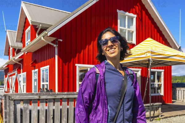 Happy tourist wearing sunglasses and purple jacket smiling in front of a vibrant red building in tofino, a popular tourist destination on vancouver island, british columbia, canada, during summer