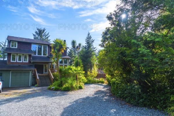 Lush green vegetation surrounds a charming two story house with a gravel driveway and attached garage in tofino, british columbia, on a sunny summer day