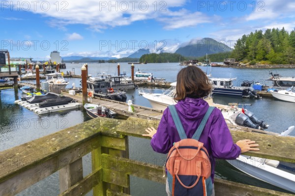 Young woman with a backpack enjoying the scenic view of boats in tofino harbor on a sunny summer day, framed by blue skies and forested mountains