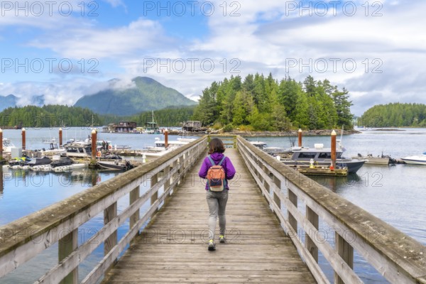 Female tourist with backpack walking down a wooden pier in tofino, british columbia, with boats, harbor, and forested island in background on a sunny summer day