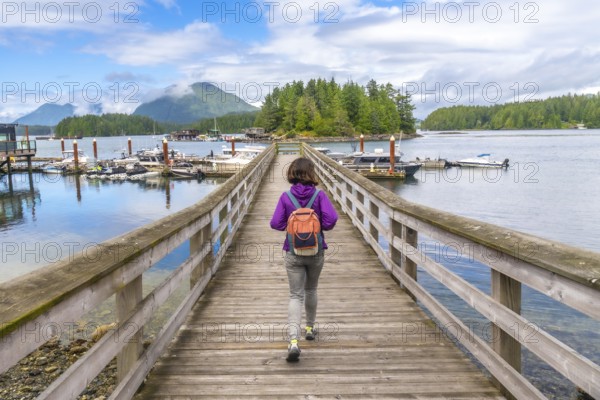 Female tourist walking down a wooden pier in tofino, british columbia, with boats and lush islands in the background during a sunny summer day