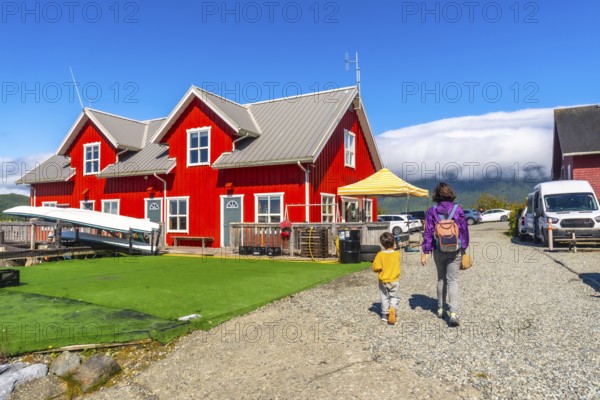 Mother and son are walking on a gravel path near a vibrant red house with white trim, canoes resting on a green turf under a clear blue sky in tofino, vancouver island
