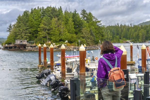 Tourist in a purple jacket and backpack capturing photos of the scenic tofino harbor, featuring moored boats and a forested island backdrop