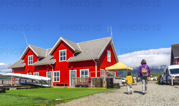 Mother and son walking on gravel path near vibrant red wooden building with canoes, enjoying summer day in tofino, vancouver island, british columbia, canada