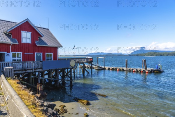 Calm, clear blue water lapping against the shore complements a vibrant red wooden building and pier in tofino, british columbia, with distant mountains and a small wind turbine enhancing the scene