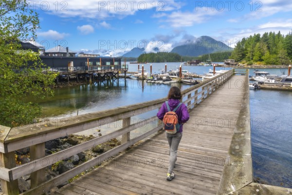 Female tourist with backpack walking down a wooden pier in tofino, british columbia, canada, enjoying the view of moored boats and lush rainforest mountains