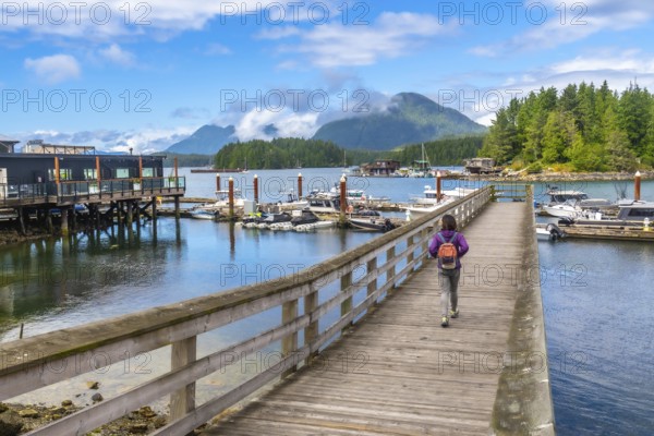 Tourist with backpack walking down a pier in tofino, british columbia on a beautiful summer day with boats, mountains, and blue sky in the background