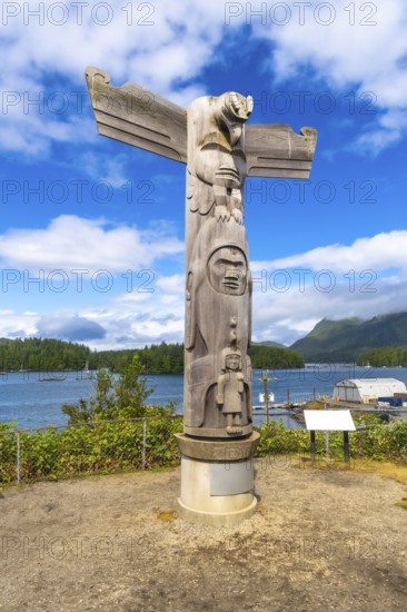 Wooden totem pole featuring intricate carvings of a thunderbird and other figures, rising against a blue sky, white clouds, and tofino's scenic waterfront in british columbia