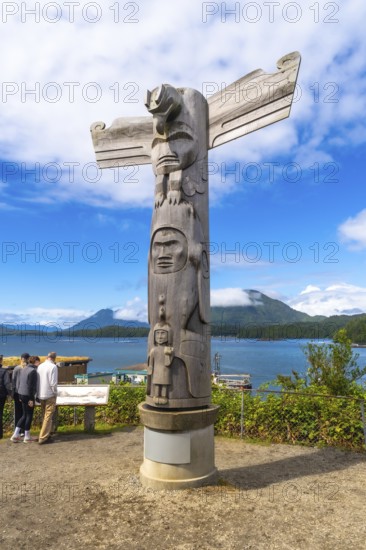 Wooden totem pole standing on a base overlooking the harbor in tofino, british columbia, canada, with tourists observing a map in the distance