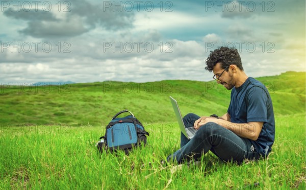 Smiling freelancer young man with laptop working from the field. Close up of man sitting on green grass working with his laptop. Smiling man with laptop sitting on the grass in the field