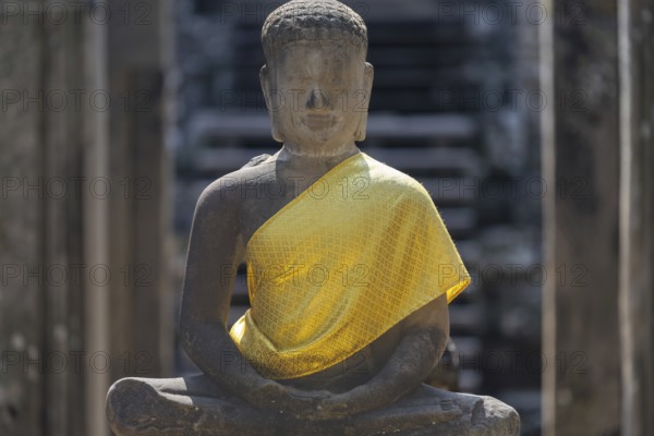 Sandstone Buddha statue, (Dhyana-mudra: gesture of meditation), with yellow tunic, on the north side of the Bayon Temple, with huge stone-carved faces of the Bodhisattva Lokeshvara, also known as Avalokiteshvara, Angkor Thom, UNESCO World Heritage Site, Angkor Wat, Siem Reap, Cambodia