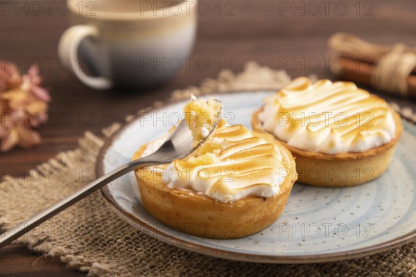 French lemon tart with meringue on brown wooden background, cup of coffee, linen textile, side view, close up, selective focus