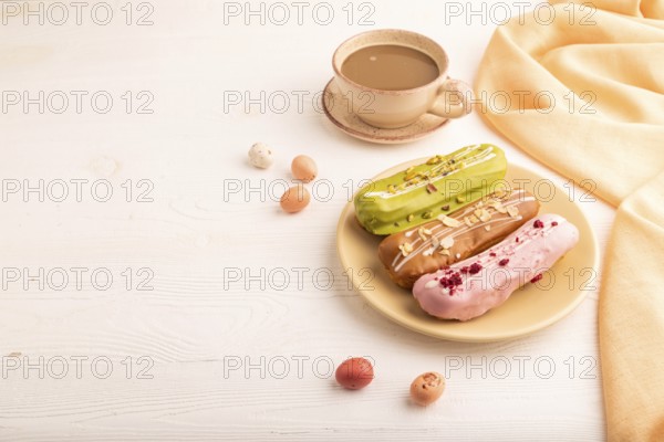 Glazed Eclairs on white wooden background and orange linen textile, cup of coffee, side view, copy space