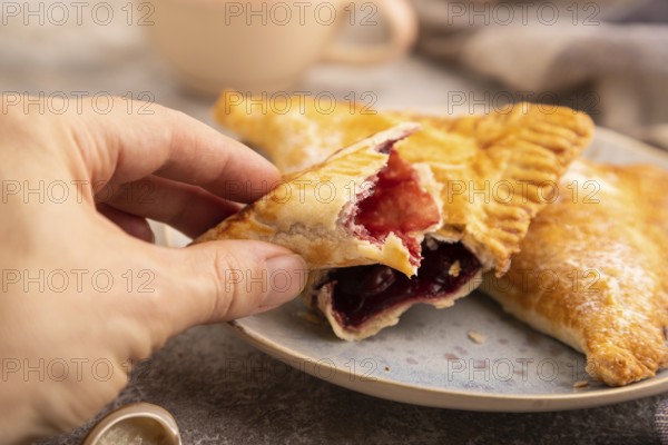 Triangles Pie, Puff Pastry, Samsa with cherry jam on brown concrete background and linen textile with hand, cup of coffee, side view, close up, selective focus