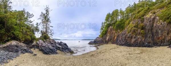 Panoramic view of a secluded beach and inlet in tofino, vancouver island, featuring dramatic rock formations, lush green vegetation, and a lone kayaker enjoying the serene waters under a cloudy sky