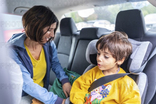 Smiling mother fastening a seat belt for her son in a child safety seat inside a car, ensuring his security and comfort during their travel together