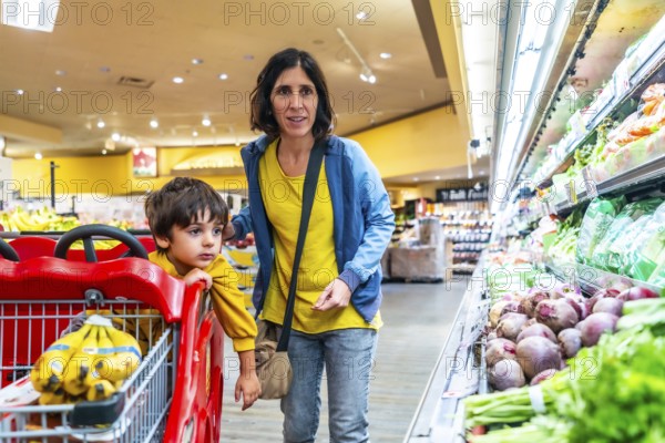 Mother and son shopping together for fresh produce in a supermarket, selecting healthy food options for their family dinner while pushing a cart down the colorful refrigerated aisle