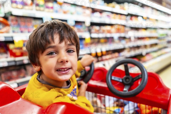 Cheerful child is enjoying a shopping trip, sitting in a shopping cart with a toy steering wheel and smiling, surrounded by colorful grocery store shelves