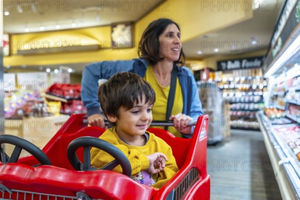 Mother pushing a shopping cart with her son sitting inside, enjoying their grocery shopping experience together in a brightly lit supermarket