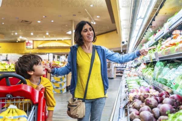 Mother points to fresh vegetables while shopping with her son in the produce section of a grocery store, promoting healthy eating habits and family bonding