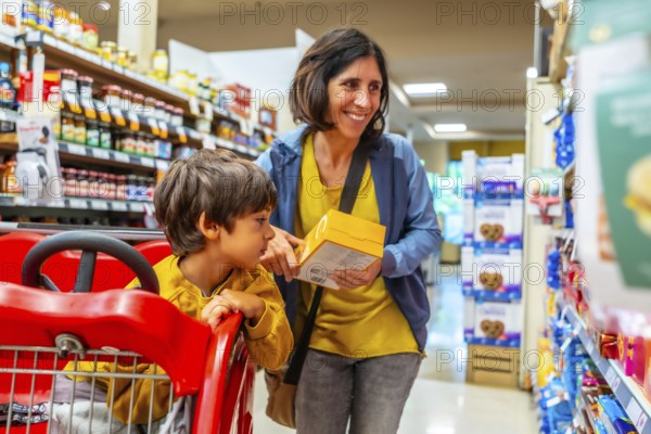 Smiling mother holding a yellow box while shopping with her son sitting in a shopping cart in a supermarket aisle, choosing products together