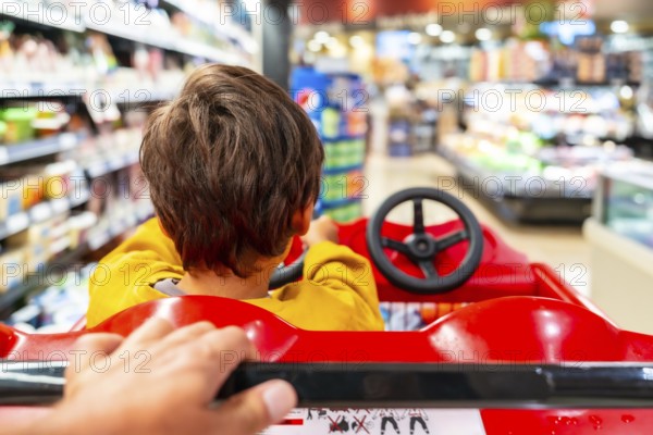 Parent pushes a shopping cart designed as a toy car, with their child pretending to drive, navigating through the aisles of a supermarket, creating a fun and engaging shopping experience