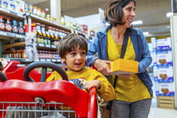 Mother holding a box of cookies next to her son sitting in a shopping cart, enjoying their grocery shopping experience together in a brightly lit supermarket