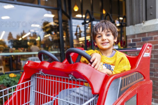 Smiling preschooler having fun sitting in a special children's shopping cart shaped like a firetruck, enjoying a shopping trip with family outside a grocery store