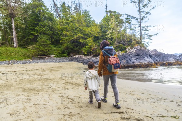 Mother and son enjoying a leisurely stroll along a beautiful sandy beach in tofino, british columbia, surrounded by lush green forest and rocky coastline, creating a serene and picturesque scene