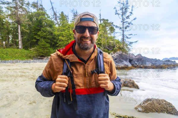 Happy tourist wearing sunglasses and baseball cap is holding his backpack straps while enjoying a sunny day at mackenzie beach in tofino, known for its stunning natural beauty and surfing culture