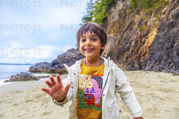 Happy child waving on a sandy beach with rocky cliffs and ocean in the background, enjoying a sunny day in tofino, vancouver island, british columbia, canada