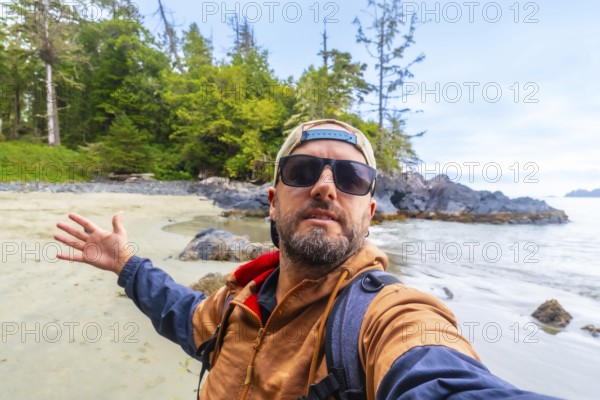 Tourist with sunglasses and baseball cap taking a selfie on chesterman beach, showing the beautiful landscape of tofino on vancouver island, british columbia, canada, during a sunny day