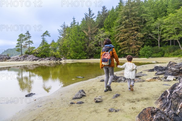 Mother and child enjoying a leisurely stroll along a sandy beach in tofino, british columbia, surrounded by lush forests and calm reflective waters
