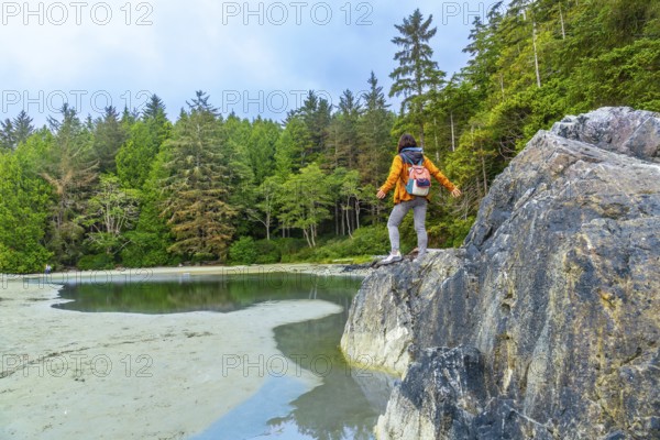 Female hiker with a backpack standing on rocks with outstretched arms, savoring the breathtaking view of mackenzie beach and the surrounding lush forest on vancouver island