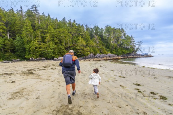 Father and son are running on a pristine sandy beach with lush green forest in the background, enjoying quality time together in the beautiful natural setting of tofino, vancouver island