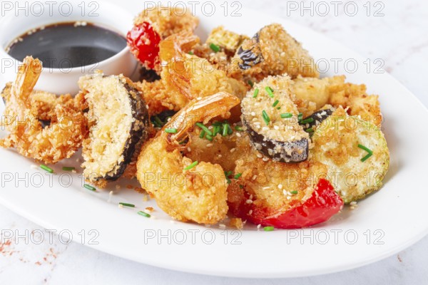 Shrimp and assorted vegetables are deep fried in a light batter and served on a white plate. A small bowl of soy sauce is beside the dish