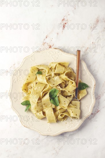 Fresh papardelle pasta, with basil and pesto sauce, is served, on a white plate, with a fork held by hand