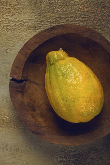 A vibrant yellow fruit, possibly a lemon or a ripe papaya, sits in a handcrafted wooden bowl against a textured background. Natural light highlights its glossy skin and freshness