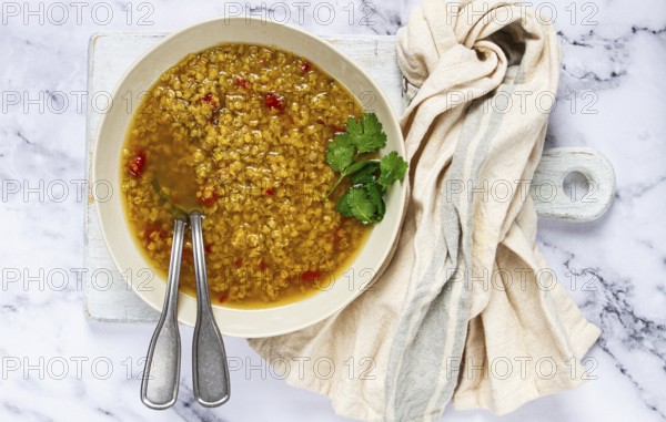 A bowl of delicious lentil soup sits on a marble countertop, garnished with fresh herbs. Silver utensils are placed beside a soft kitchen towel, creating a warm, inviting atmosphere