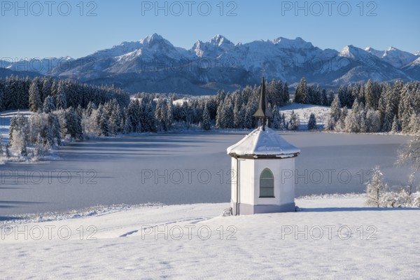 Chapel on the shore of a frozen lake surrounded by snow and mountains under a blue sky, Hegratsrieder See, near Füssen, Ostallgäu, Allgäu, Bavaria, Germany