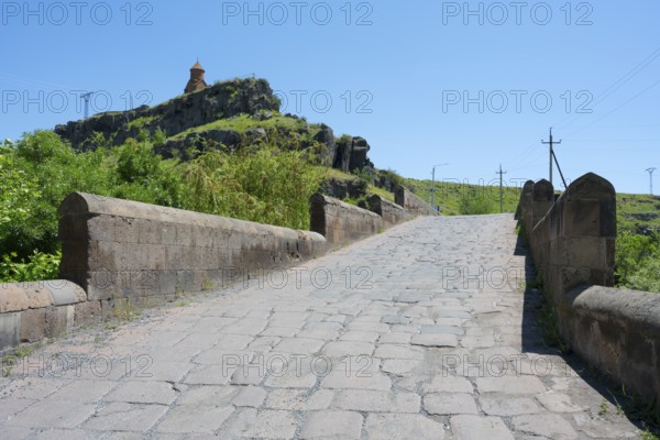 A stone bridge leads to a hill with a church under clear sky and green landscape, St. Sarkis church and three-arched bridge over Kassagh from 1664, Ashtarak bridge, Ashtarak, Aragazotn province, Armenia