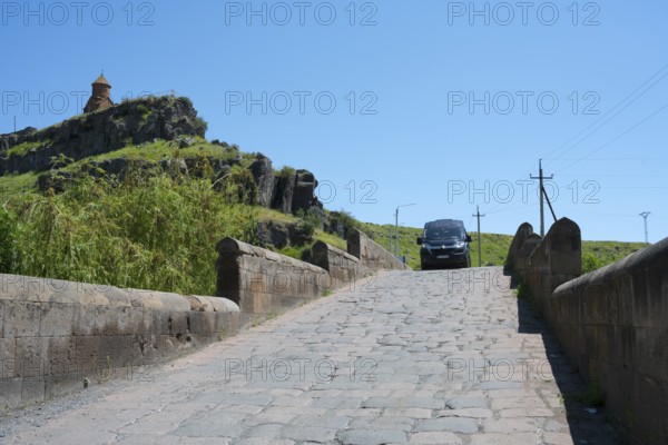 A car drives over a stone bridge with a building on a green hill in the background under clear sky, St. Sarkis church and camper on a three-arched bridge over the Kassagh from 1664, Ashtarak, Aštarak, Aragazotn province, Armenia